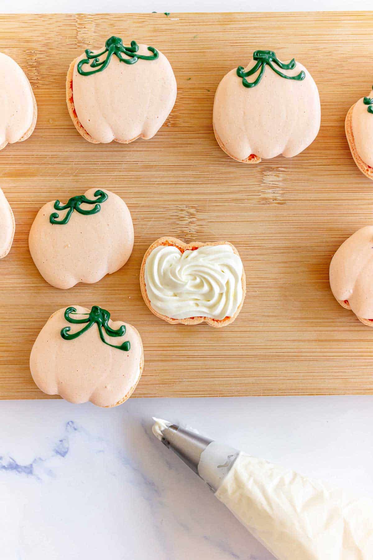 Pumpkin-shaped macarons on a wooden board, with one macaron open and filled with cream, piping bag nearby.