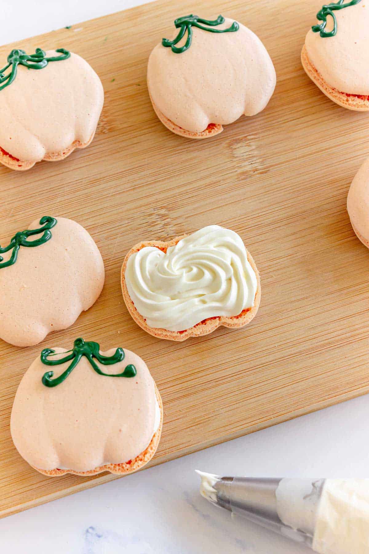 Pumpkin-shaped macarons on a wooden board, one is open and filled with white cream, piping bag nearby.