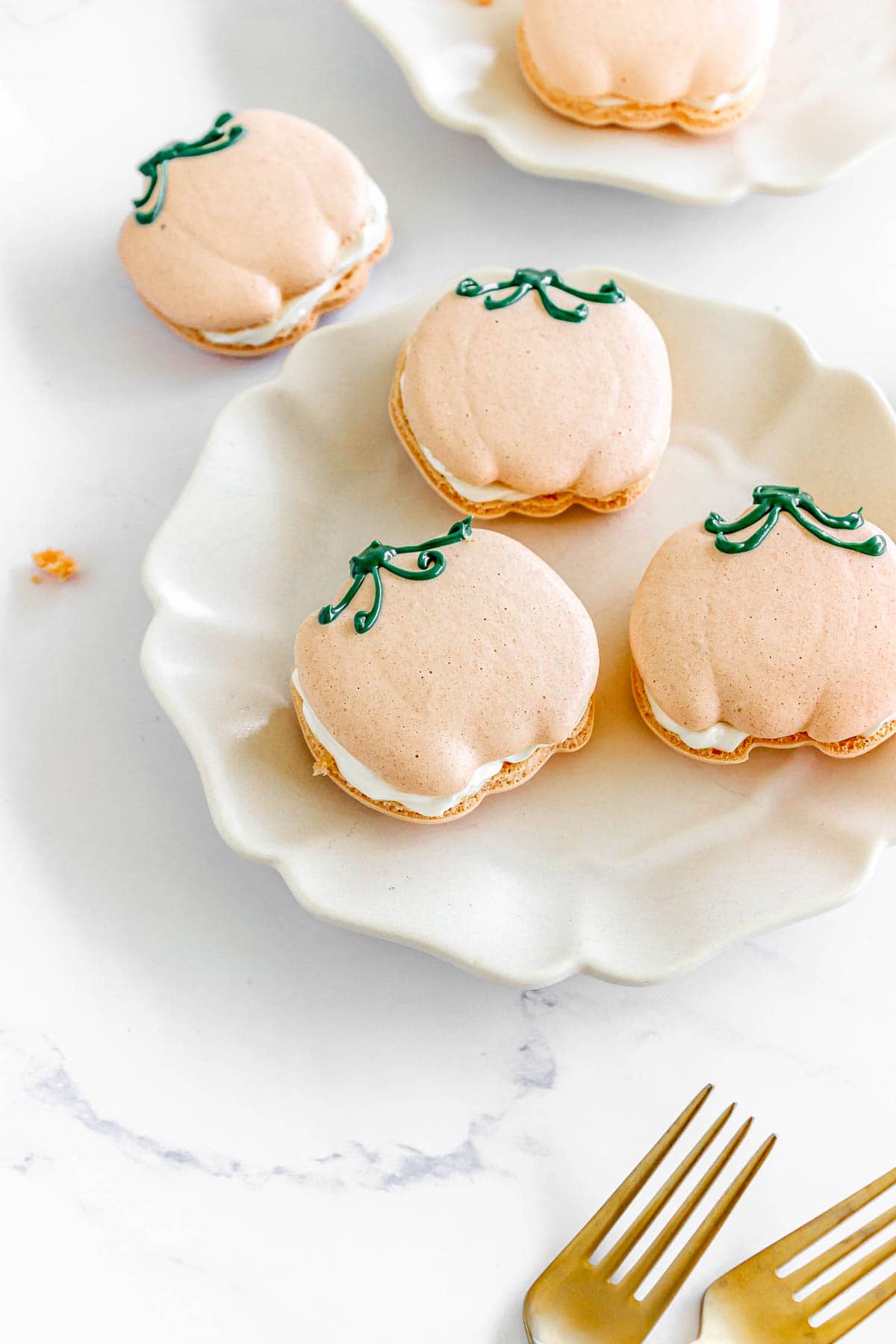 Pumpkin-shaped macarons with white filling and green icing on white plates, next to two gold forks.