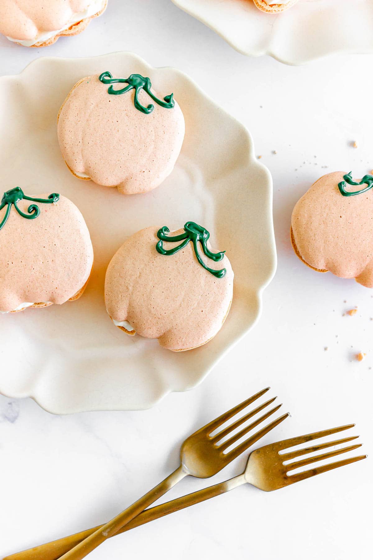 Pumpkin-shaped macarons with green icing stems on white plates, with two gold forks nearby.