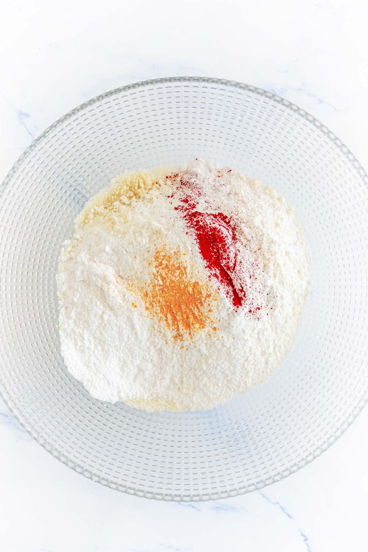 A glass bowl with flour and red and orange spices on a white surface.