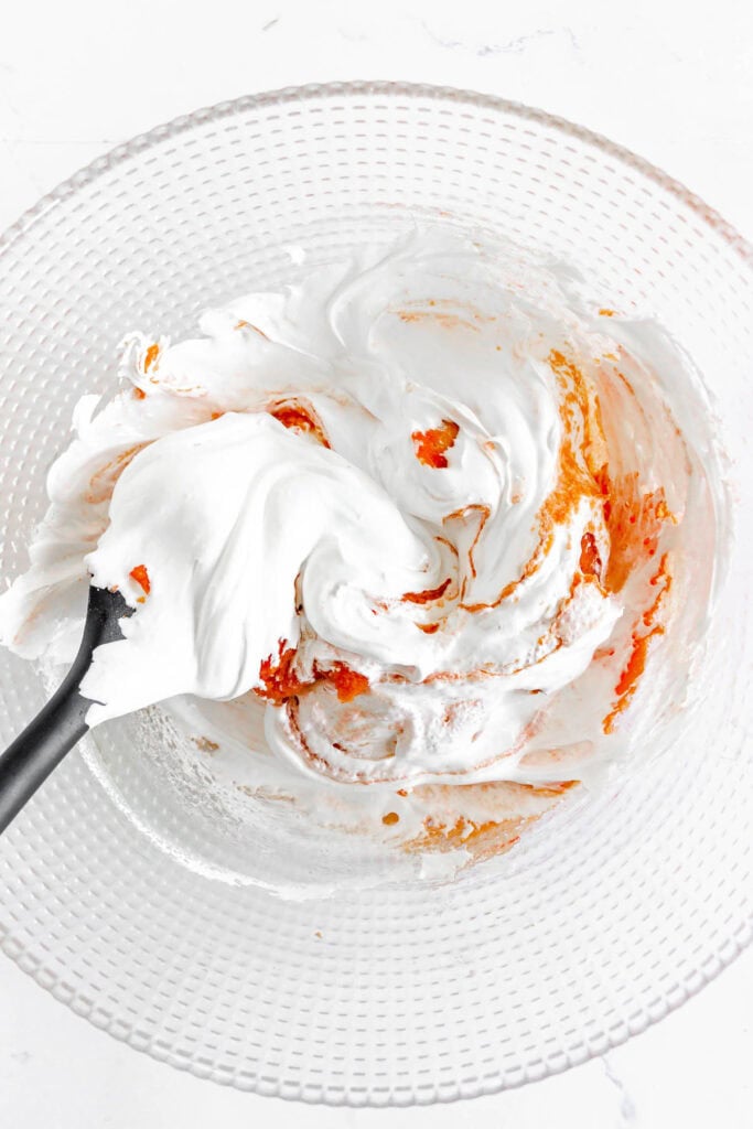 A bowl of white whipped mixture being stirred with orange food coloring and a spatula.