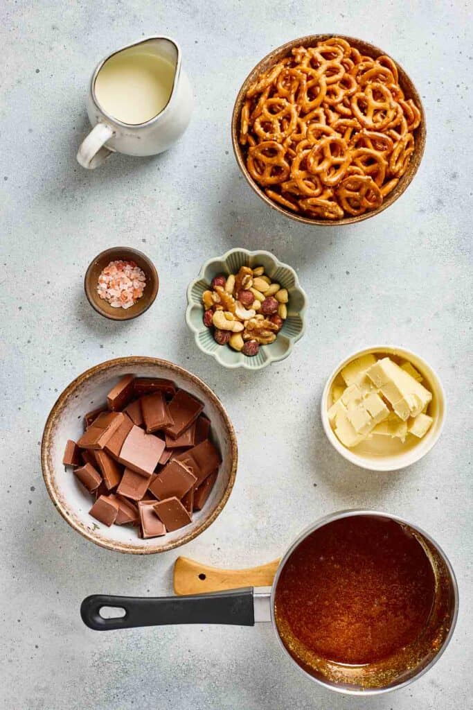 Bowls of Salted Caramel Pretzel bark, mixed nuts, chocolate, butter, a small bowl of salt, cream, and a saucepan of caramel.