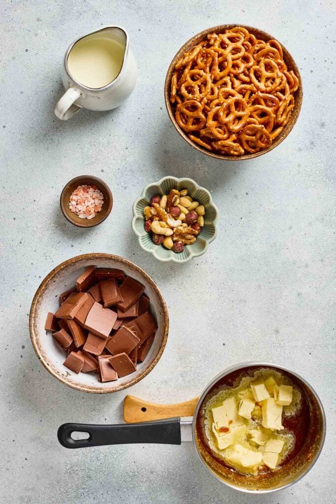 Bowls of pretzels, chocolate, mixed nuts, salt, cream, and a saucepan with melting butter on a light surface&mdash;perfect for creating salted caramel pretzel bark.