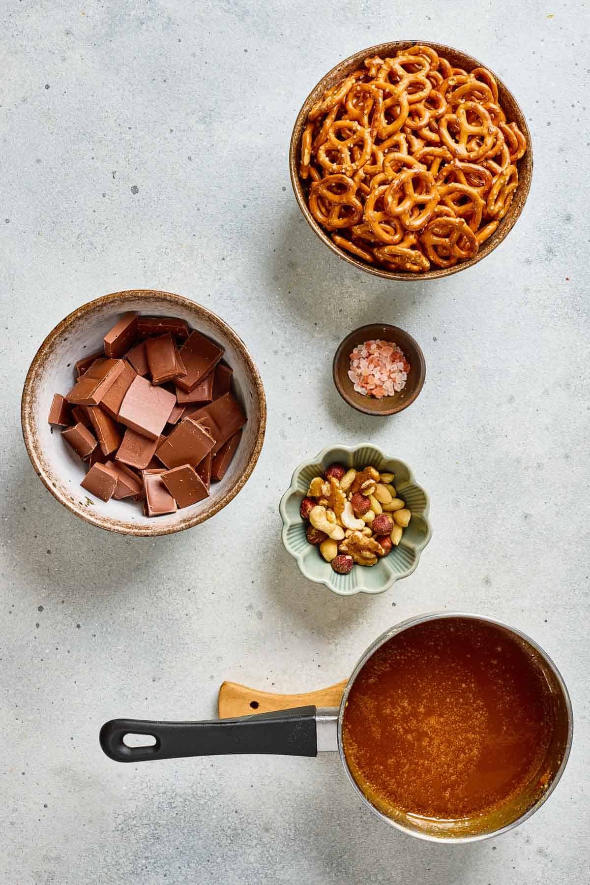Bowls of pretzels, chocolate chunks, mixed nuts, pink salt, and a saucepan of melted salted caramel create the perfect setup for making delicious Salted Caramel Pretzel Bark on your countertop.