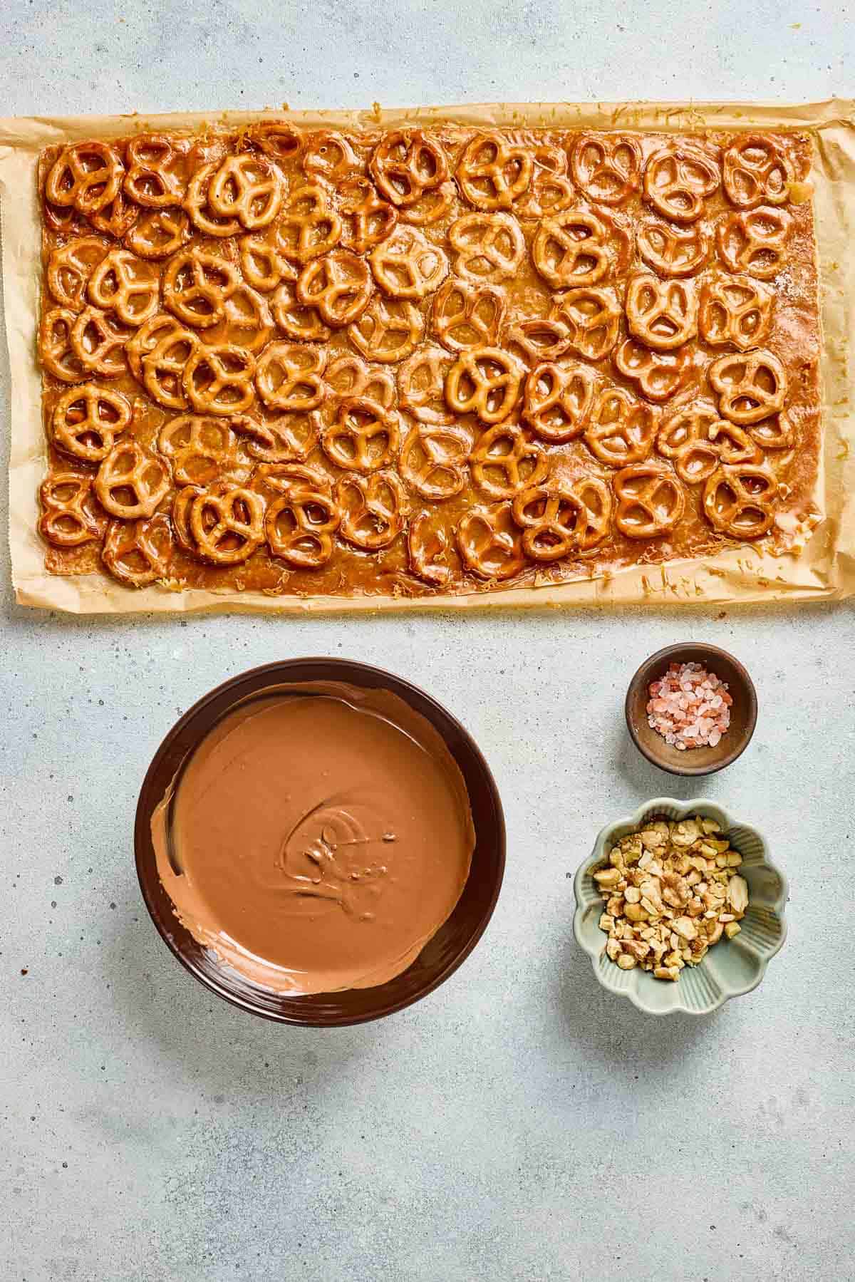 Sheet of pretzels on parchment, ready to be transformed into Pretzel Bark, with bowls of melted chocolate, chopped nuts, and pink salt nearby.