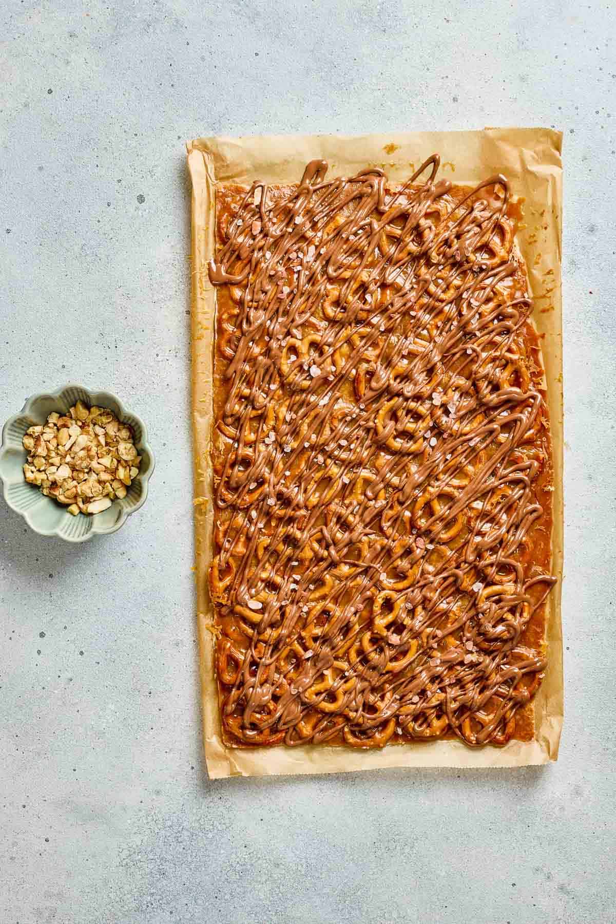 Rectangular sheet of salted caramel pretzel bark topped with crunchy pretzels and chocolate drizzle on parchment, next to a bowl of mixed nuts.