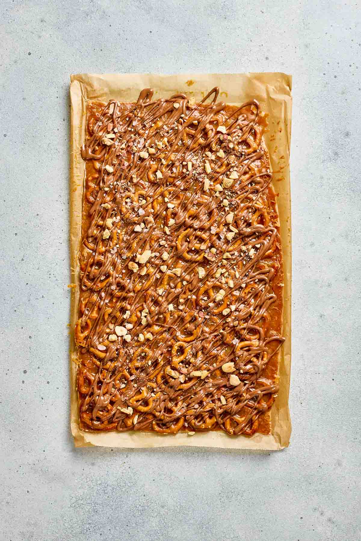 Rectangular slab of salted caramel pretzel bark topped with pretzels, chopped nuts, and chocolate drizzle on parchment paper.