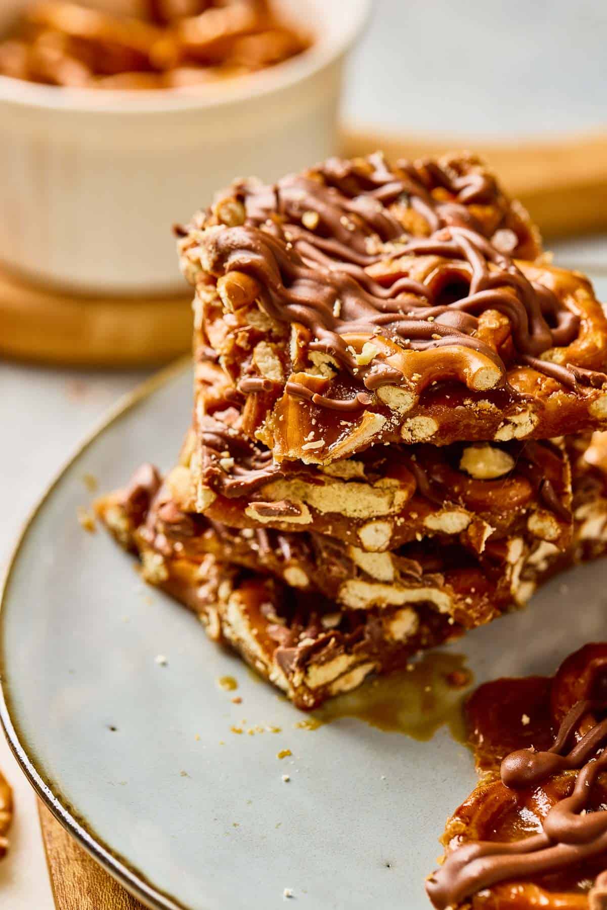 A stack of salted caramel pretzel bars drizzled with chocolate sits on a plate, with a bowl in the background&mdash;perfect for fans of pretzel bark.