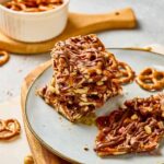 Stacked caramel pretzel bars drizzled with chocolate on a plate, with pieces of salted caramel and pretzel bark scattered around.