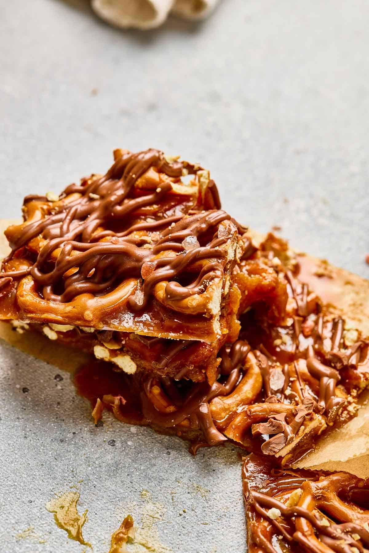 Close-up of salted caramel and chocolate-drizzled dessert bars stacked on a gray surface, reminiscent of classic salted caramel pretzel bark.