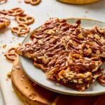 Salted Caramel Pretzel Bark pieces on a plate, drizzled with chocolate, with scattered pretzels and a ramekin in the background.
