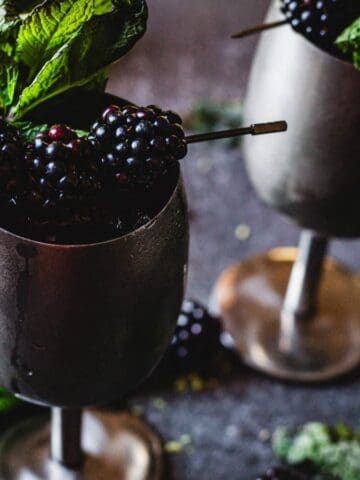 Two metal goblets filled with blackberries, garnished with mint leaves, on a table with scattered mint.