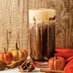Iced pumpkin spice coffee with cream, inspired by Starbucks Copycat Pumpkin Cream Cold Brew, surrounded by mini pumpkins, cinnamon sticks, and orange napkins.