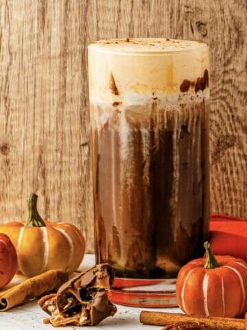 Iced pumpkin spice coffee with cream, inspired by Starbucks Copycat Pumpkin Cream Cold Brew, surrounded by mini pumpkins, cinnamon sticks, and orange napkins.