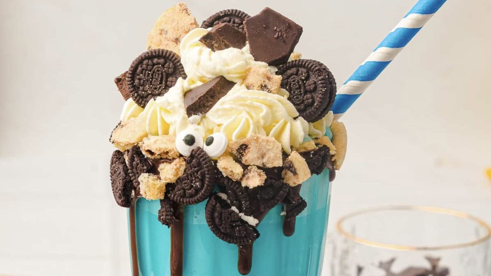 A blue cup filled with a milkshake, topped with whipped cream, mini chocolate sandwich cookies, cookie pieces, candy eyes, and a blue striped straw.