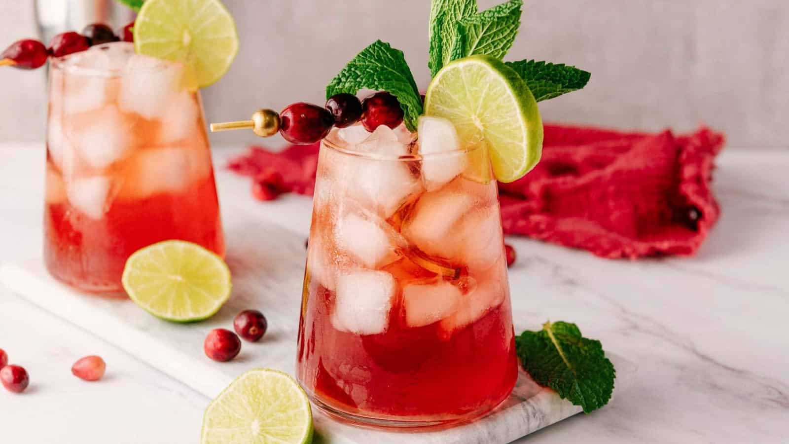 Iced cranberry drinks garnished with lime slices, cranberries, and mint on a marble board.