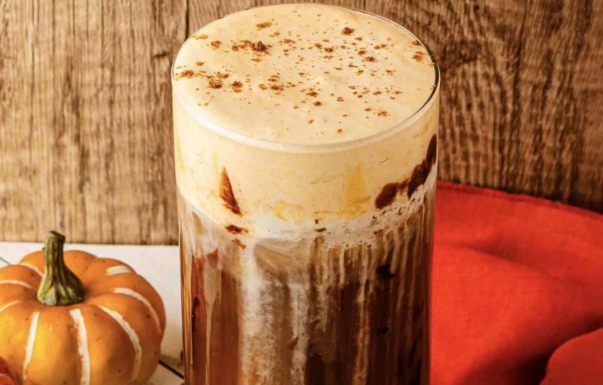 Glass of Pumpkin Cream Cold Brew topped with cinnamon, beside a small pumpkin and orange cloth&mdash;a perfect Starbucks Copycat treat for fall coffee cravings.
