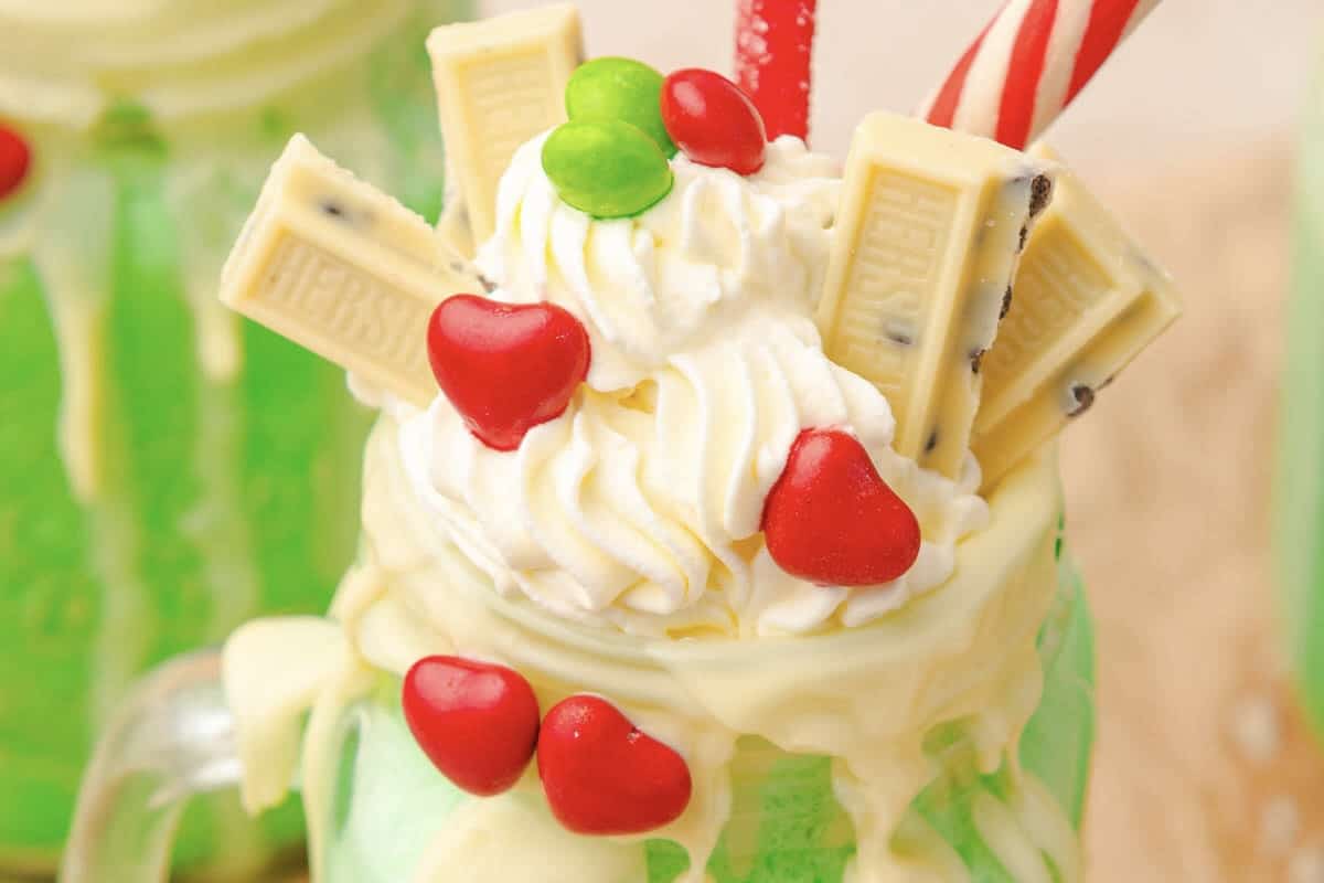 A green Grinch Freakshake topped with whipped cream, candy hearts, white chocolate, and a red-striped straw.