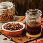 Roasted sweet toasted pecans in an orange dish and jar, with a mason jar of pecan syrup on the table.