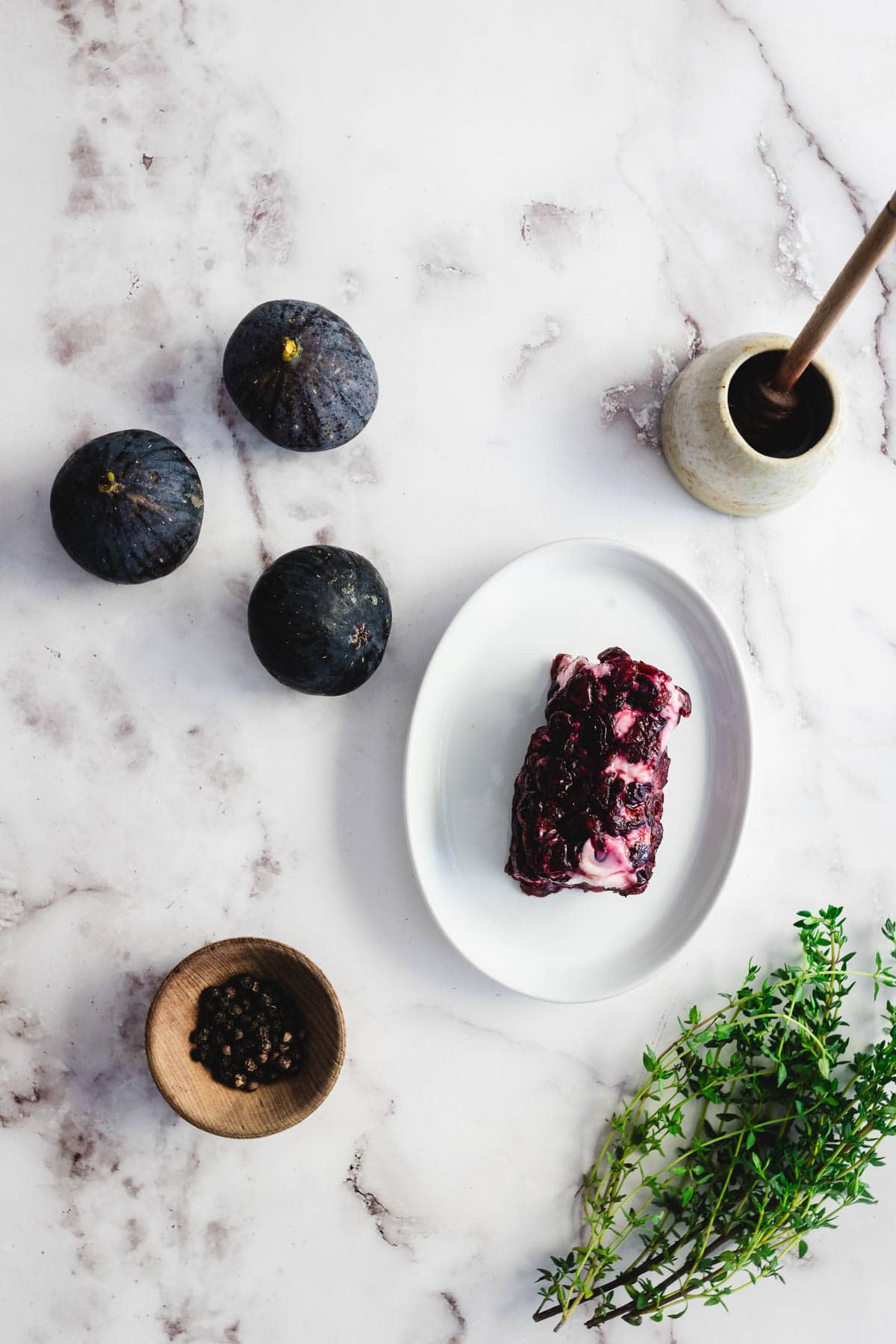 Figs, a bowl of peppercorns, herbs, and goat cheese stuffed figs are artfully arranged on a marble countertop.