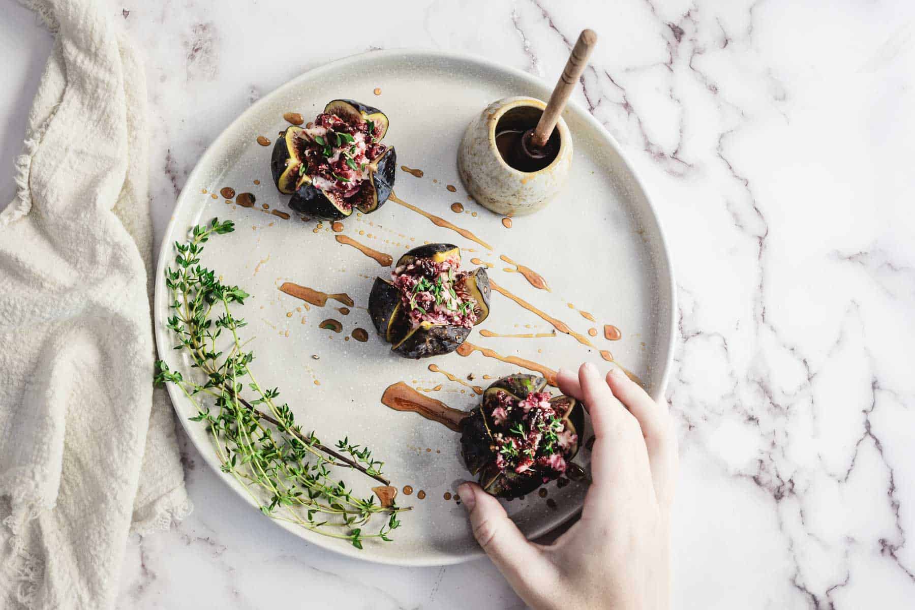 Hand reaching for Goat Cheese Stuffed Figs on a plate with sauce, fresh herbs, and a small ceramic pot on marble surface.