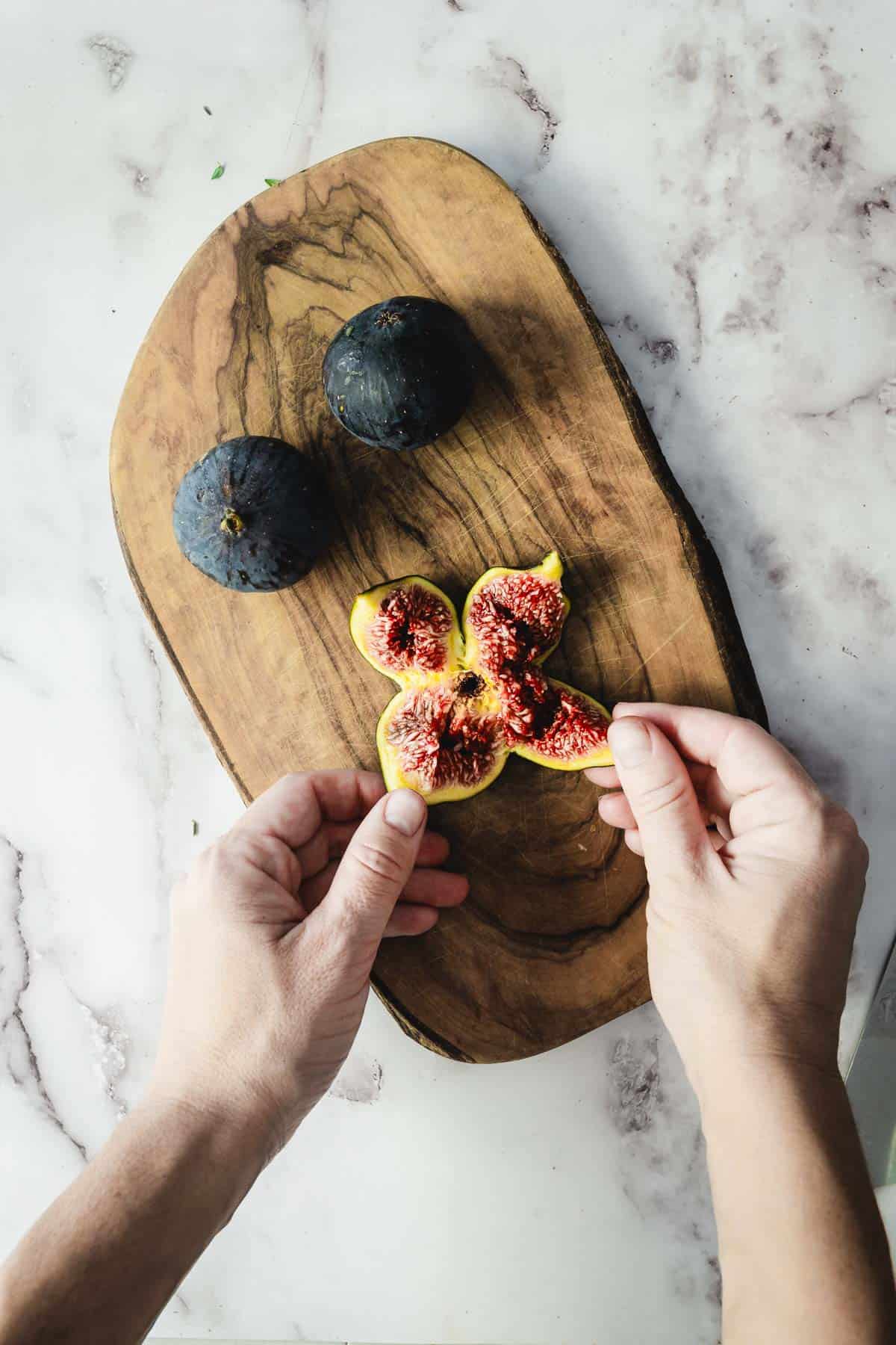 Two hands opening a ripe fig on a wooden board, perfect for making goat cheese stuffed figs, with two whole figs resting on a marble surface.