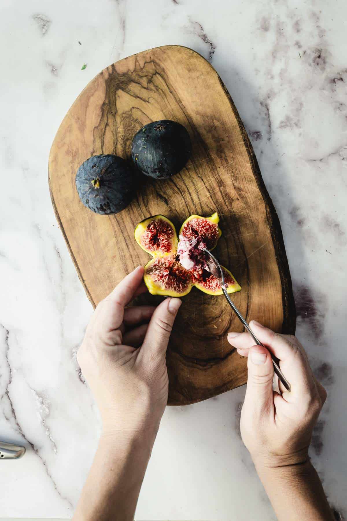 Hands scooping flesh from a halved fig on a wooden board, with two whole figs nearby&mdash;perfect for preparing Goat Cheese Stuffed Figs.