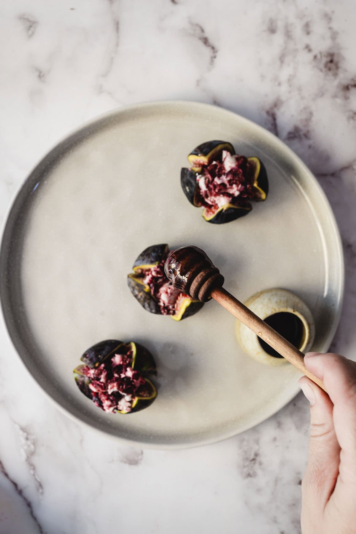 A hand drizzles honey over goat cheese stuffed figs on a round, gray plate set atop a marble surface.
