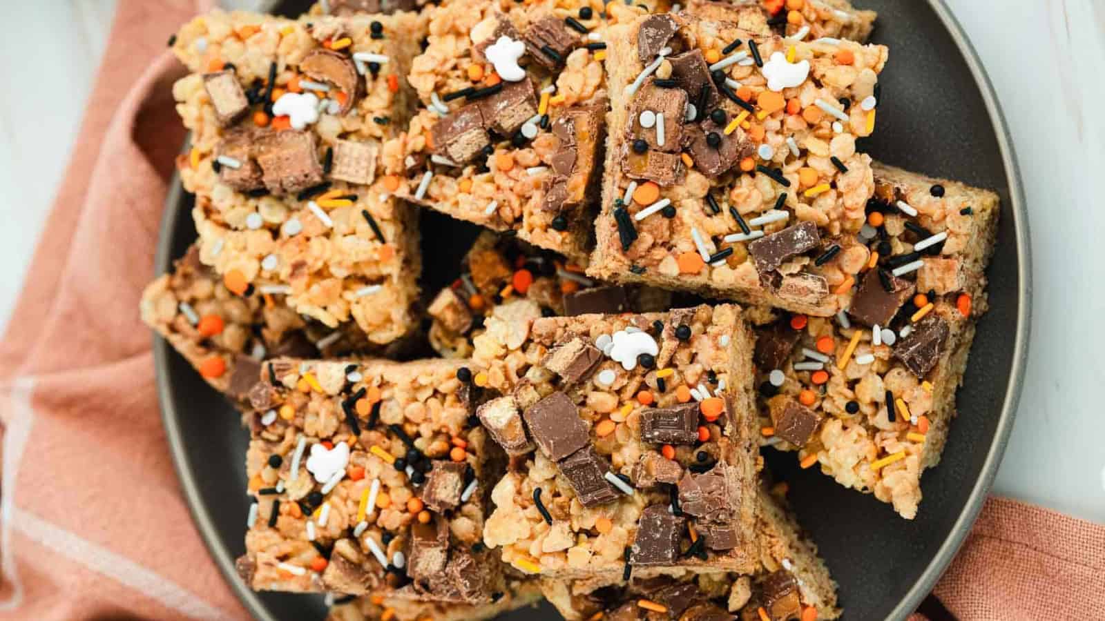 Rice crispy treats topped with chocolate pieces and colorful Halloween-themed sprinkles on a dark plate.
