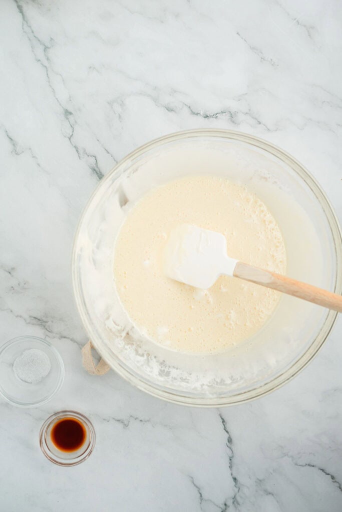 A glass bowl with creamy batter and a spatula, next to small bowls of vanilla and salt on a marble counter.