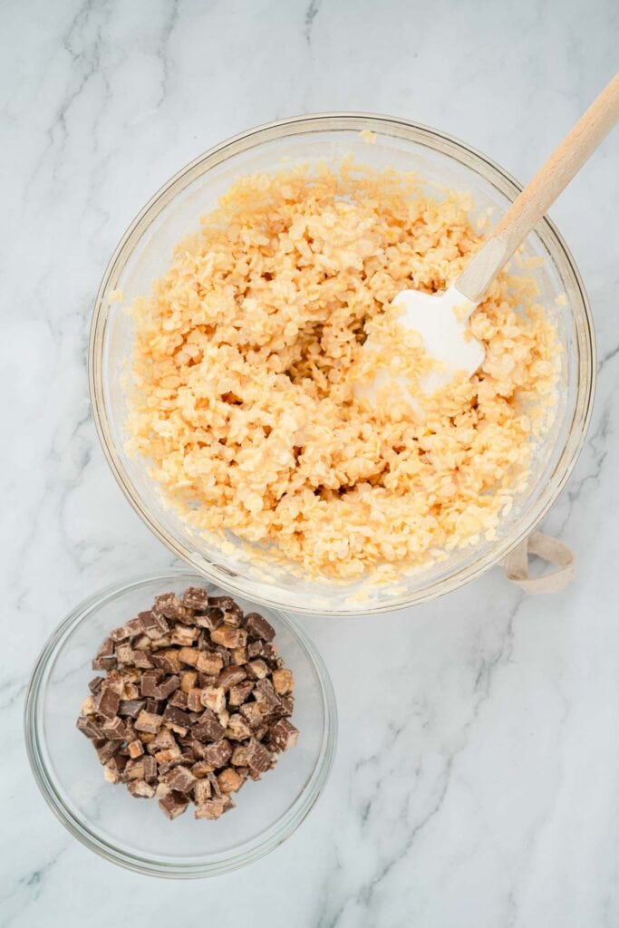 A bowl of Rice Krispies mixture with a spatula next to a bowl of chopped chocolate candy on a marble counter.
