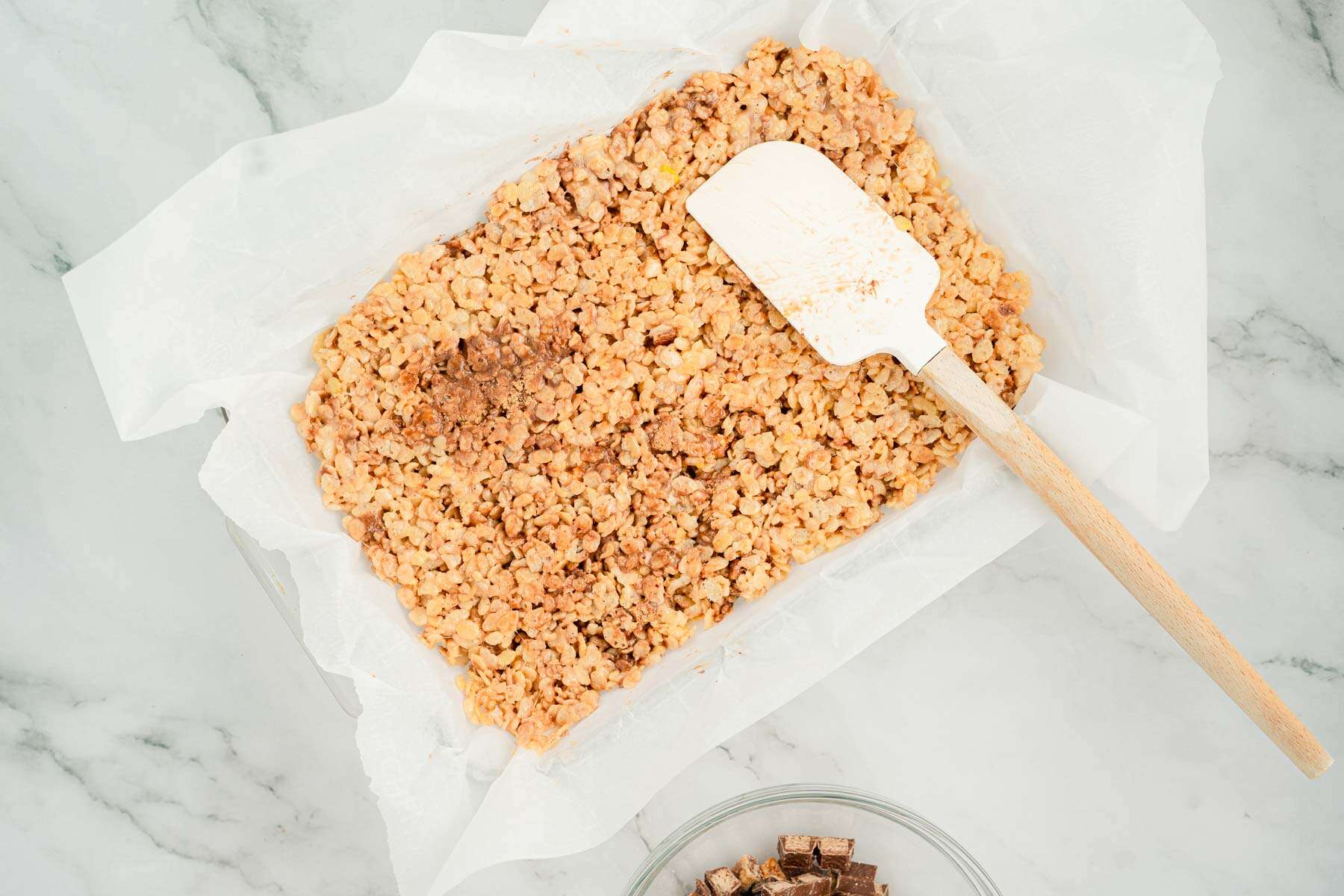 A baking pan lined with parchment paper filled with a rice crispy mixture and a spatula on top.