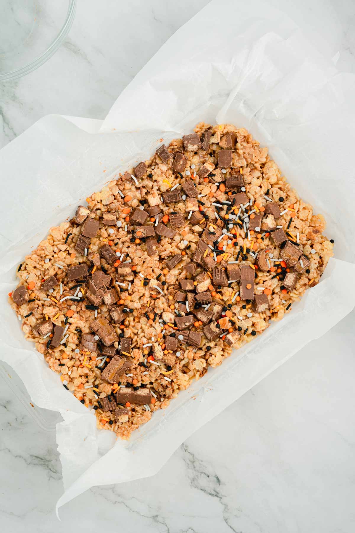 A tray of Rice Krispies treats with chocolate chunks and colorful sprinkles on parchment paper.