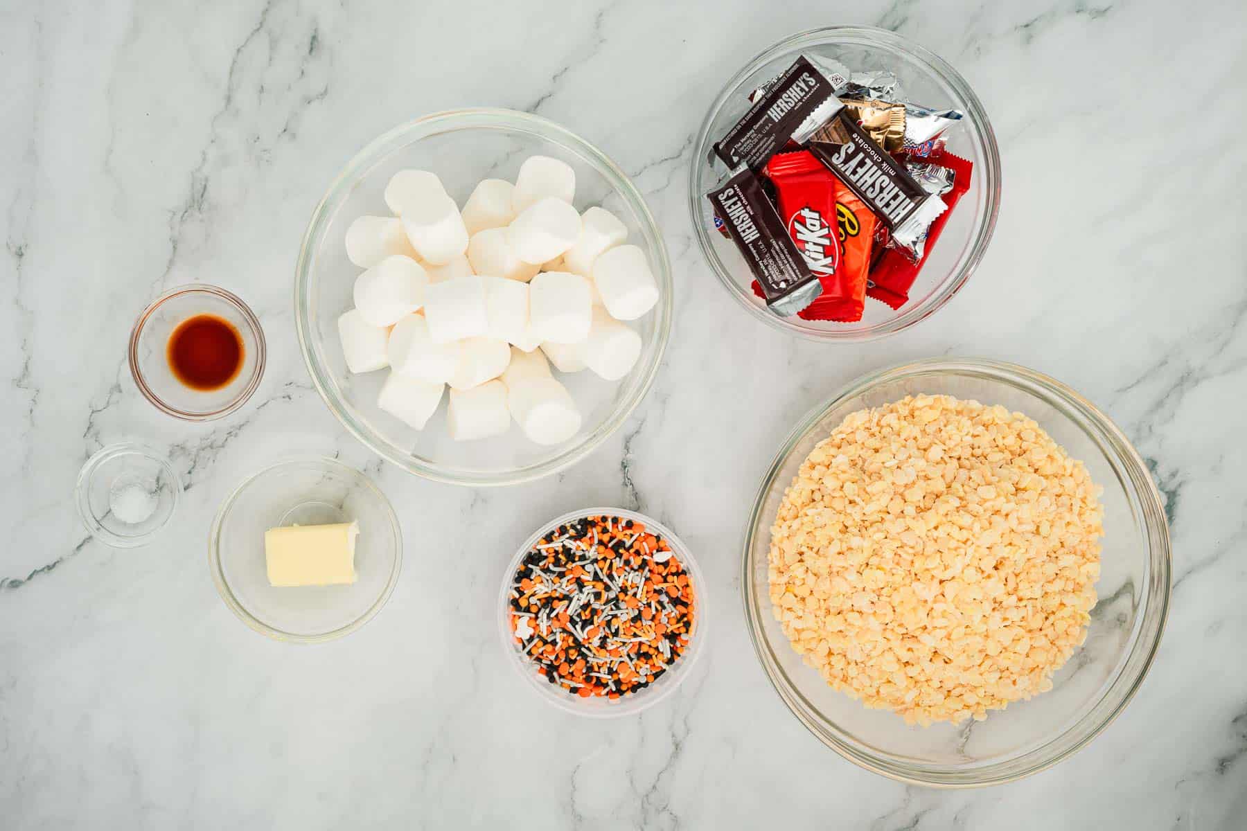 Bowls of marshmallows, rice cereal, sprinkles, butter, vanilla, and assorted candy on a marble countertop.