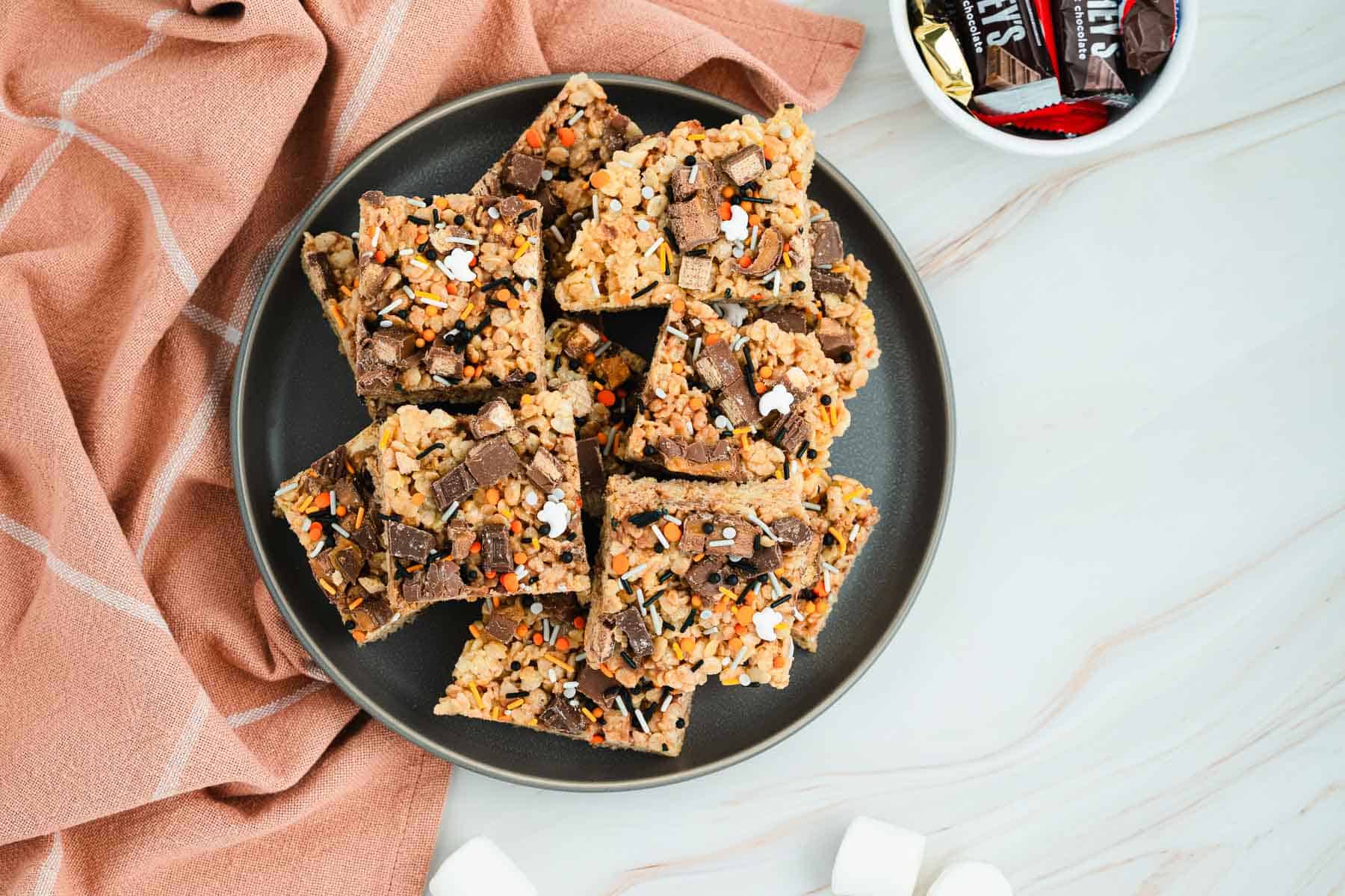 A plate of chocolate and sprinkle-topped rice crispy treats on a marble surface with a pink cloth nearby.