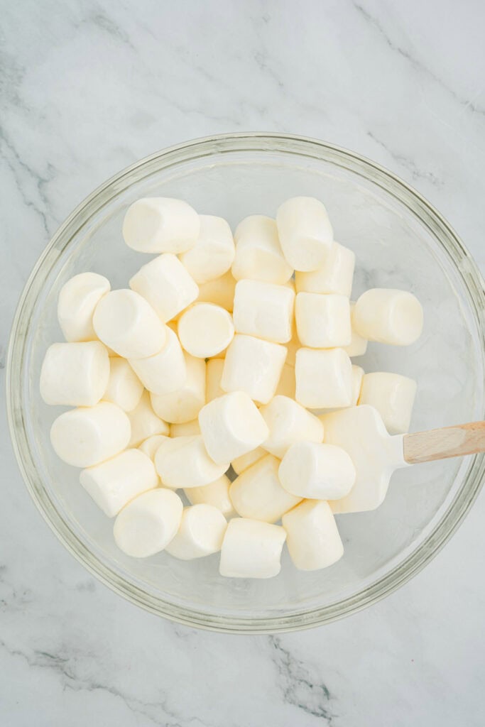 A glass bowl of white marshmallows with a white spatula on a marble surface.