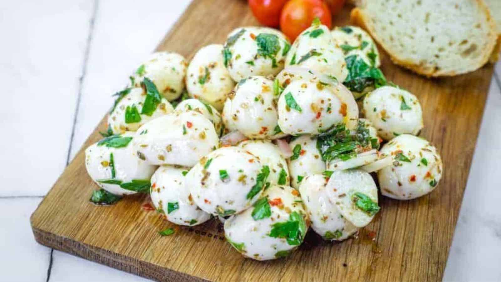 A pile of herb-marinated mozzarella balls with herbs on a wooden board, next to sliced bread and a bunch of cherry tomatoes.