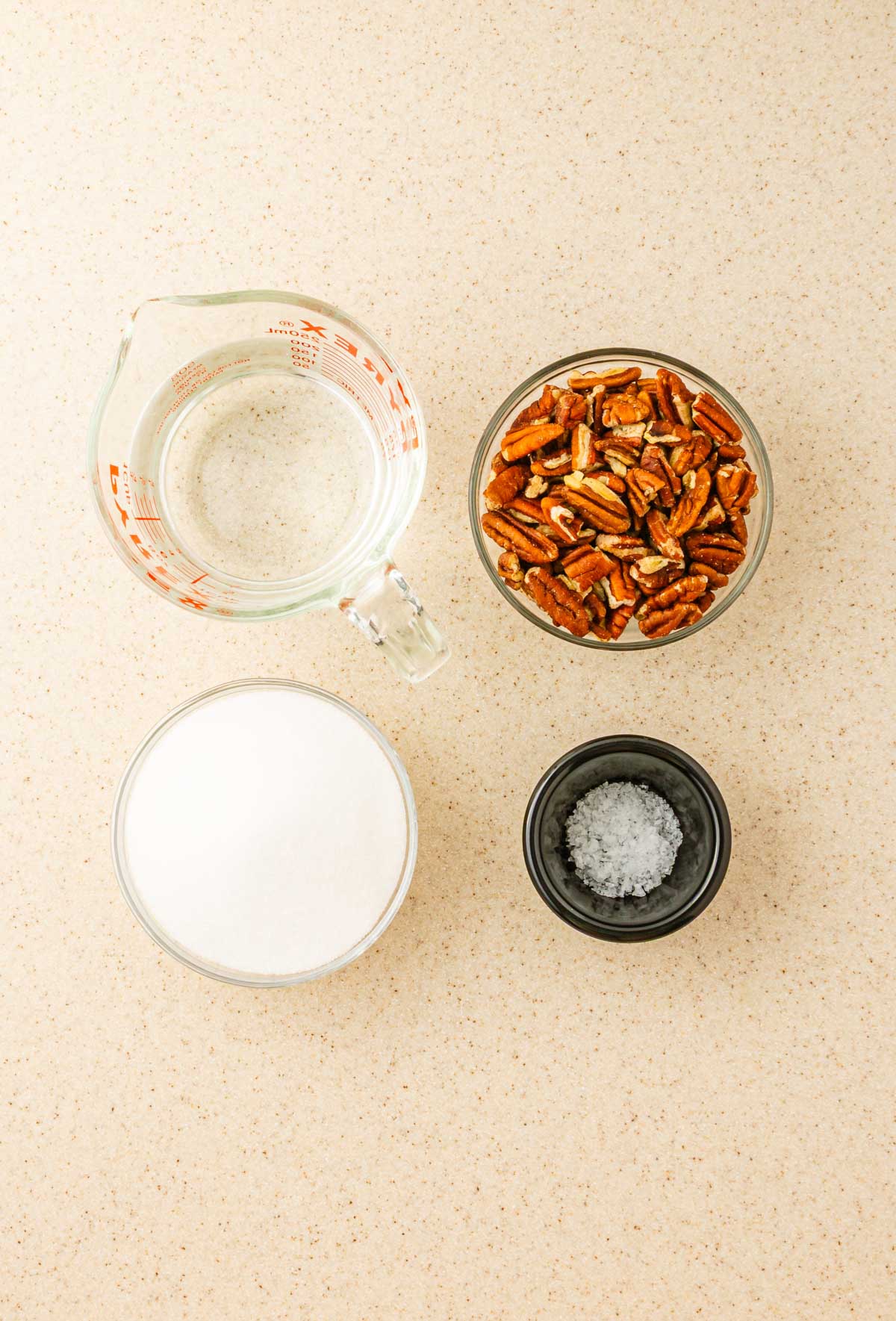 A measuring cup of water, a bowl of sugar, toasted pecans, and a small bowl of salt sit on the counter&mdash;perfect ingredients for a delicious homemade recipe.