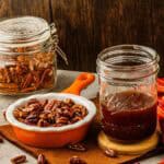 A jar of pecans, a dish of sweet toasted pecans, and a jar of homemade pecan syrup on a table with a brown napkin.