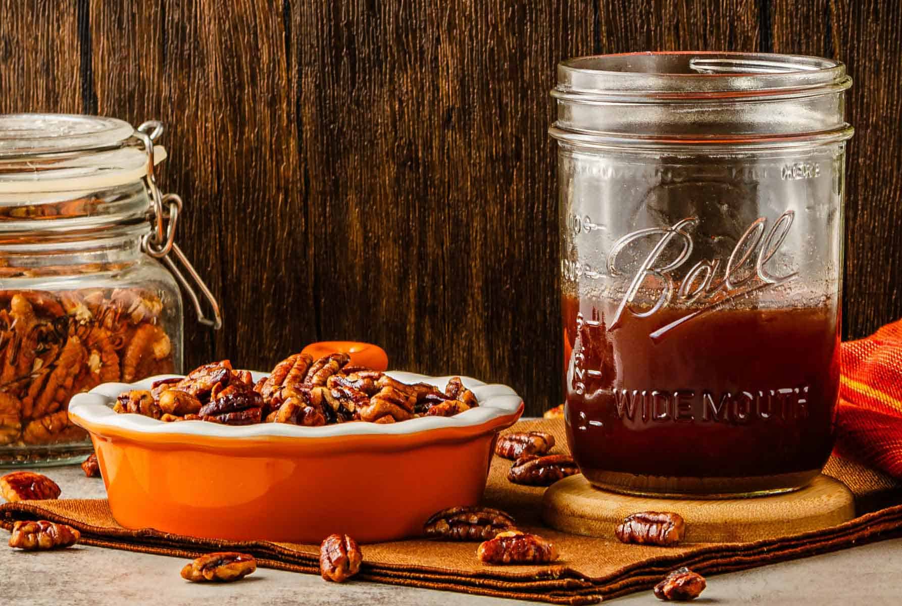 A jar of homemade pecan syrup and a dish of sweet toasted pecans rest on a table with a rustic wooden background.