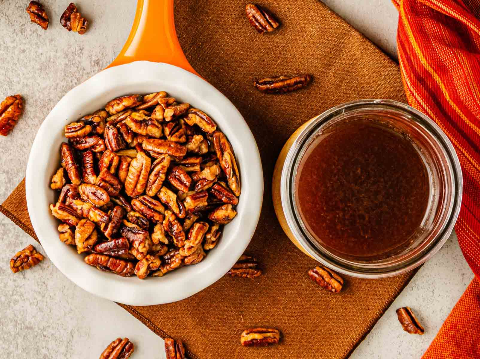 A bowl of sweet toasted pecans and a jar of pecan syrup sit on a brown napkin, with scattered pecans nearby.