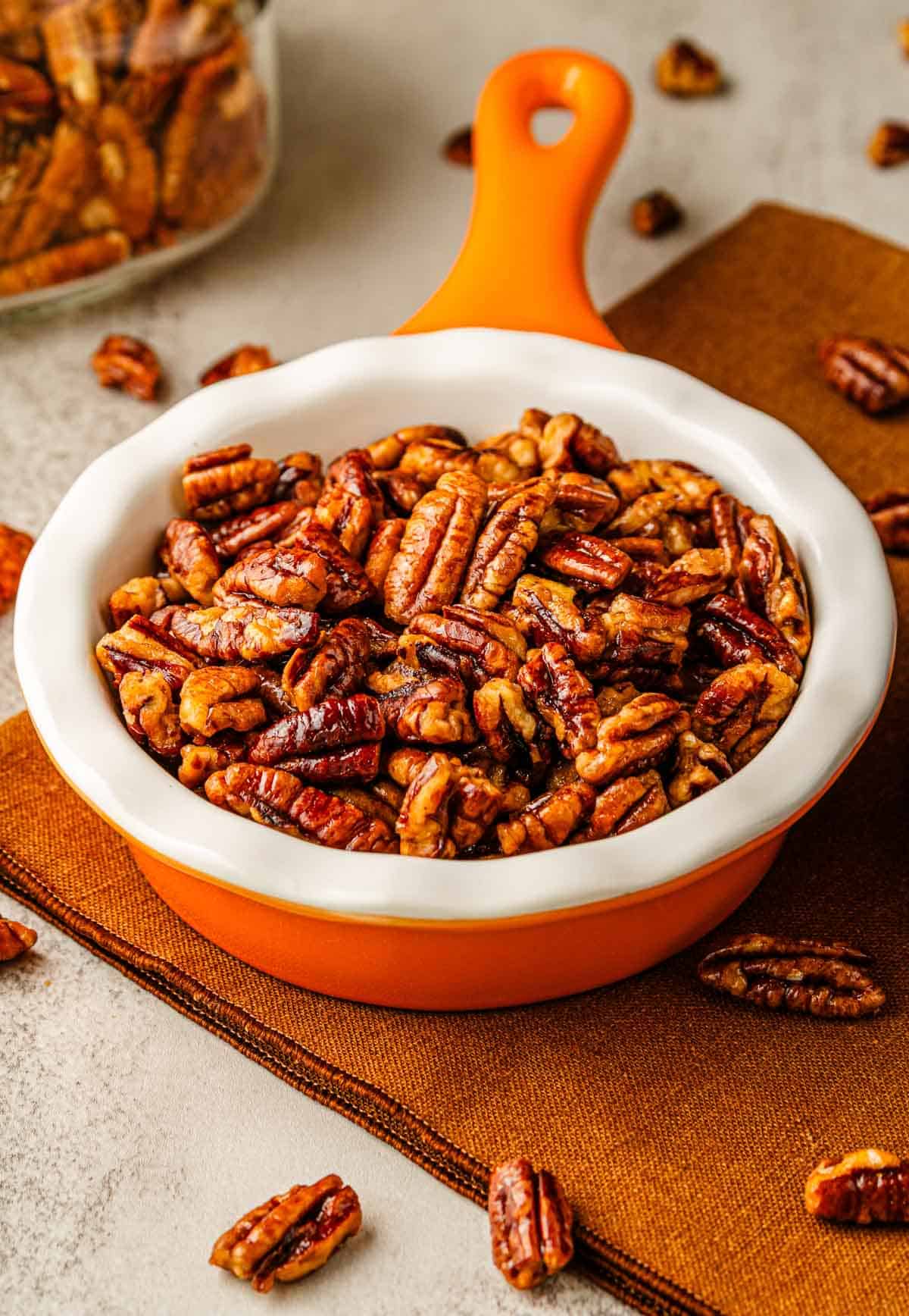 A white and orange dish filled with sweet toasted pecans sits on a brown cloth, with glazed homemade recipe pecans scattered around.