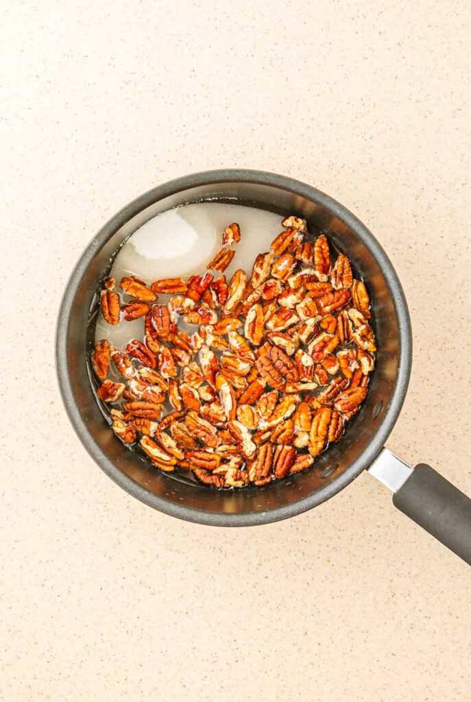Toasted pecans in sugar water for a delicious homemade recipe, simmering in a saucepan on a light countertop, viewed from above.