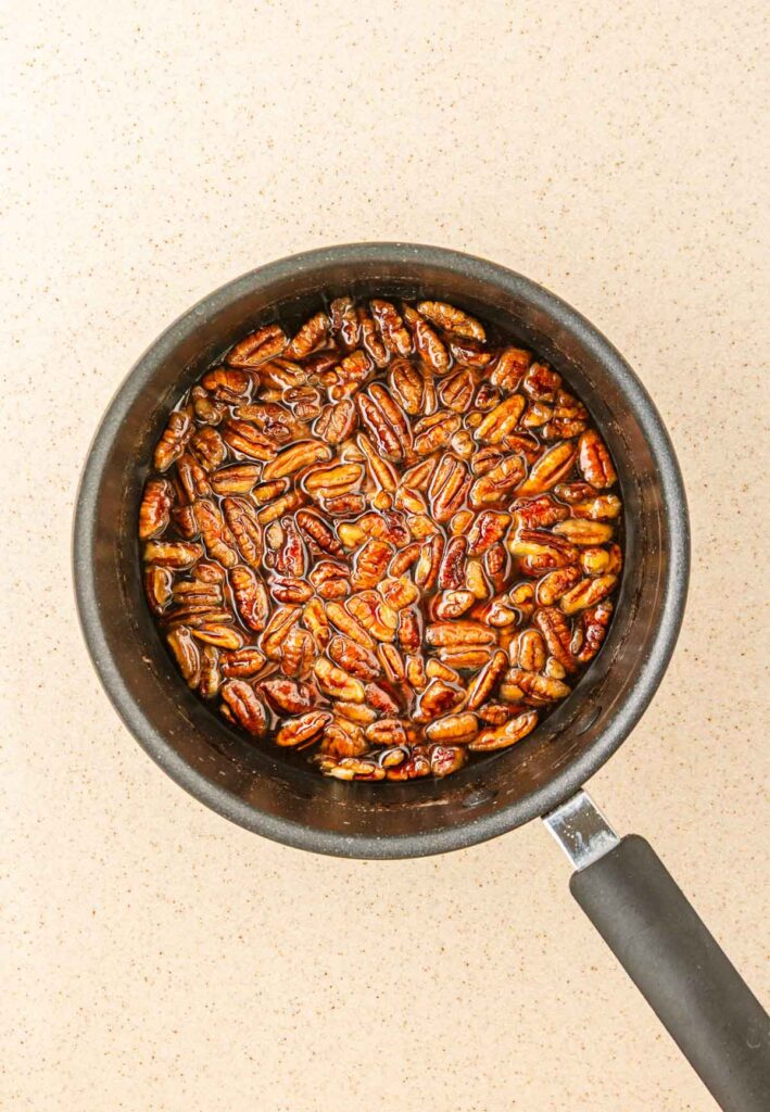 A saucepan filled with toasted pecans glazed in pecan syrup sits on a light-colored countertop, viewed from above.