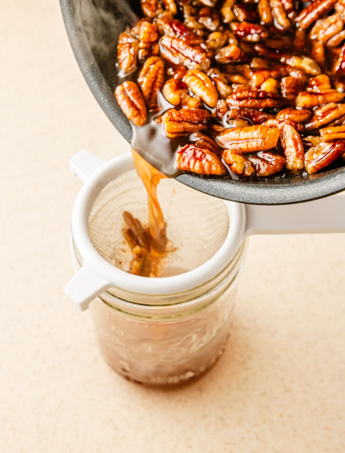 Toasted pecans and liquid being poured through a strainer into a glass jar, creating a rich pecan syrup&mdash;perfect for elevating your homemade recipe with the nutty flavor of sweet toasted pecans.