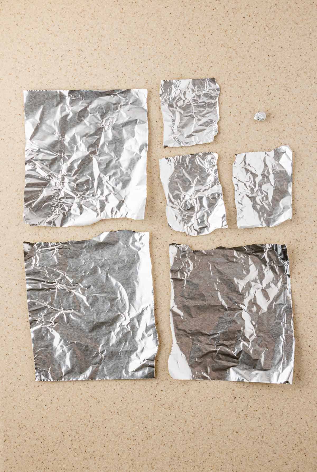 Six pieces of crumpled aluminum foil of various sizes rest on a beige countertop, as if just unwrapped from fresh Ghost Muffins or Boo-berry treats topped with cream cheese icing.