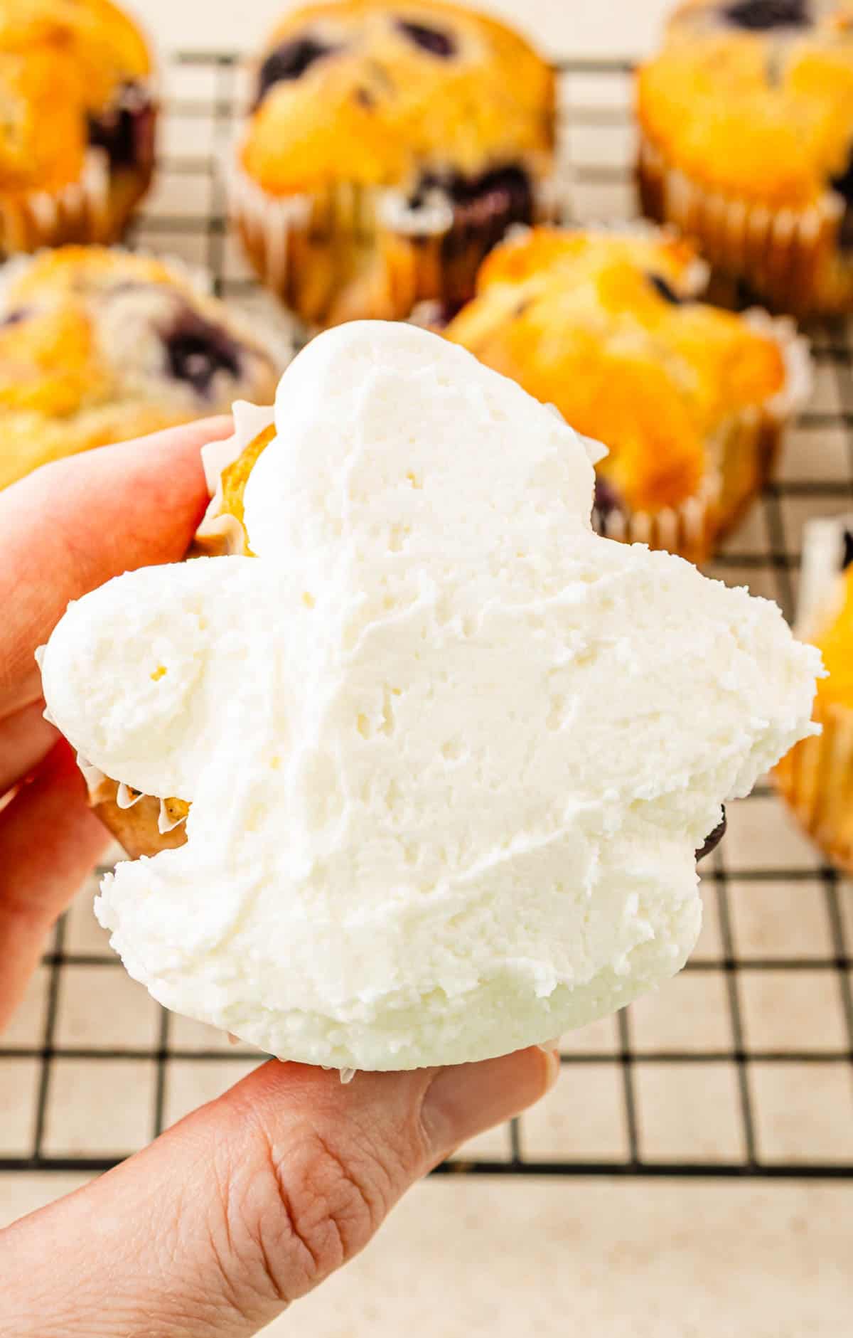 A hand holding a cupcake with white frosting and scream cheese icing; more ghost muffins are on a cooling rack in the background.