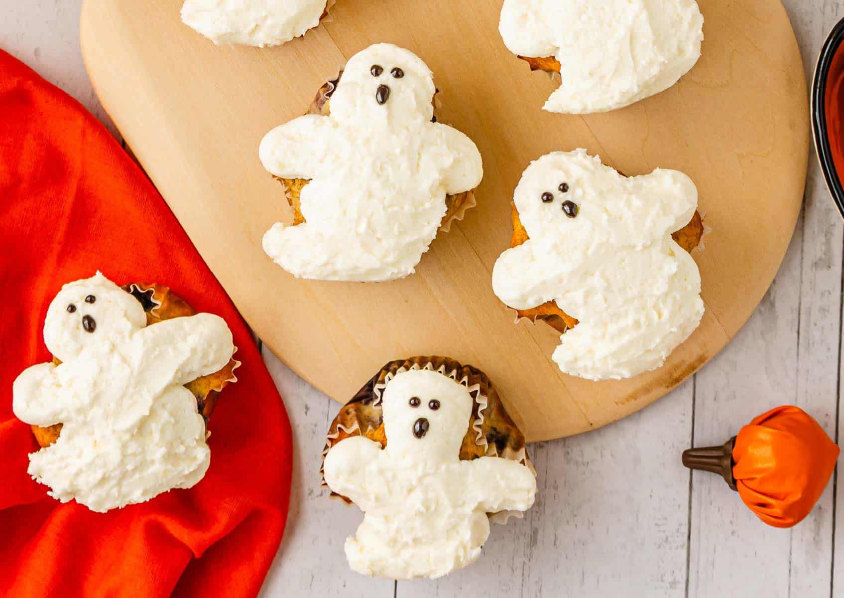 Ghost Muffins topped with SCREAM Cheese Icing, shaped like spooky ghosts, are arranged on a wooden board with a red cloth nearby.