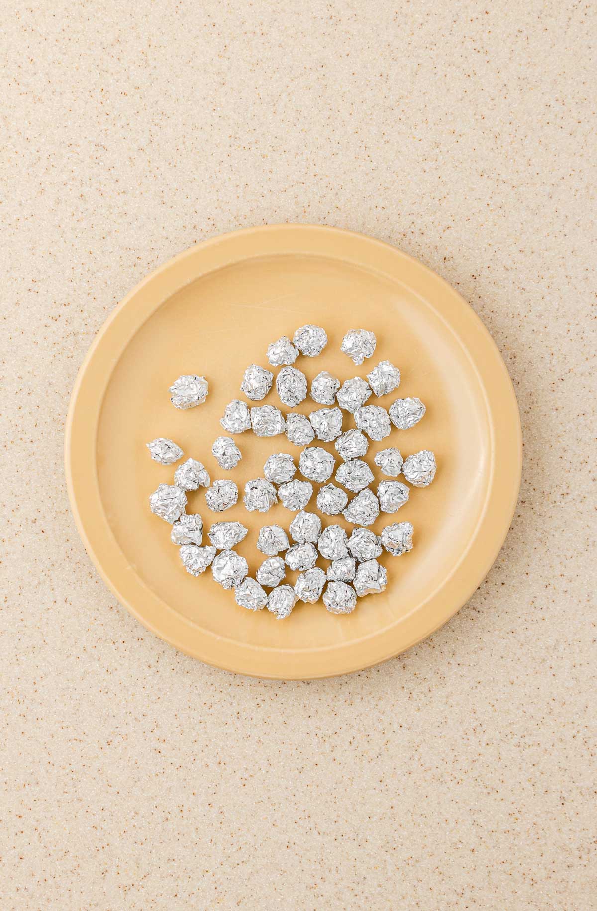 Round tan plate with several pieces of crumpled aluminum foil on a speckled beige surface, like leftover wrappers from Ghost Muffins or treats topped with Scream Cheese Icing.